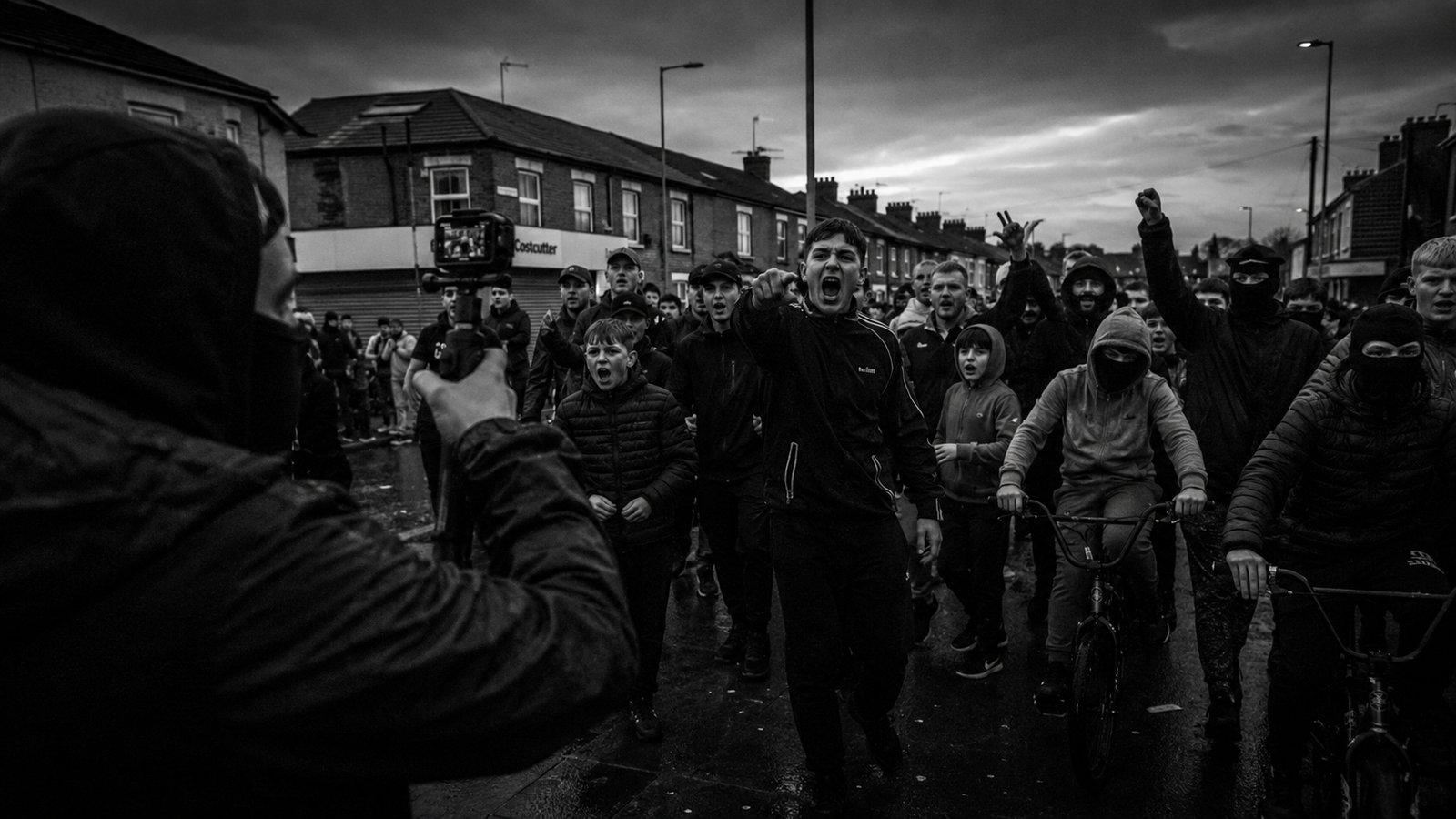 A masked crowd marches down a terraced street towards a camera